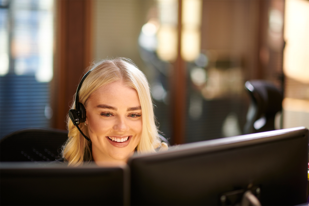 A woman on a computer with two monitors and a headset smiling.
